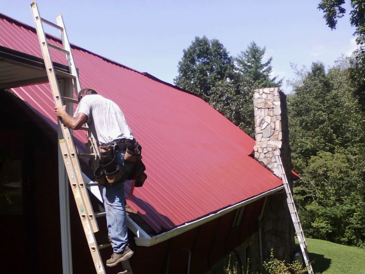 Roofer on a ladder beside a red metal roof and stone chimney — Soffit & Fascia Repair by Roofing Brothers in Baxter